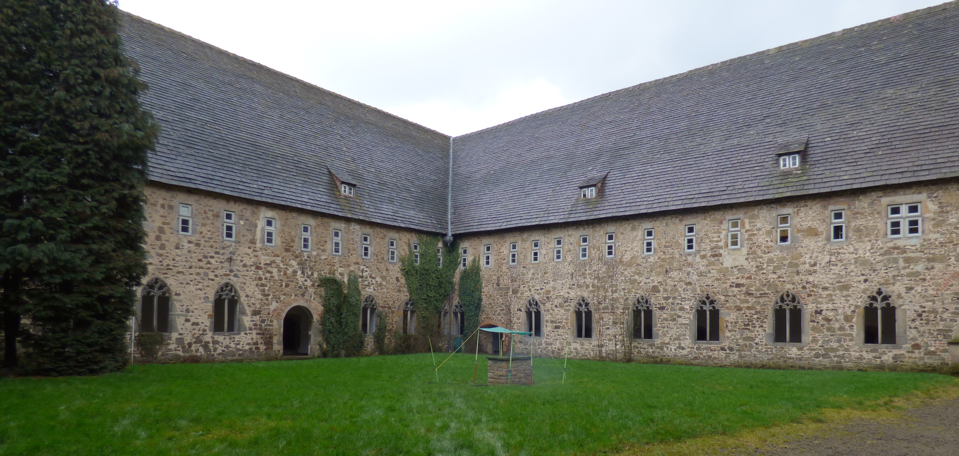 Innenhof eines historischen Geb&auml;udes mit Steinmauern, Fenstern und gr&uuml;nem Rasen, umgeben von B&auml;umen und einem tr&uuml;ben Himmel.