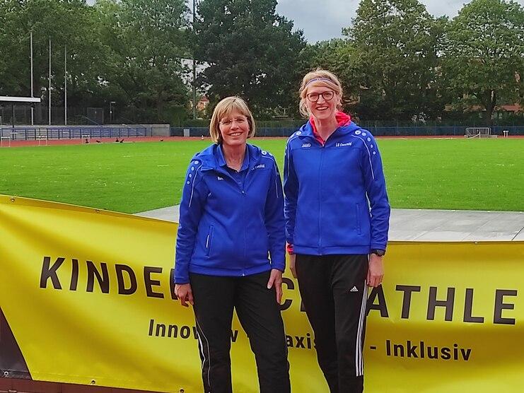 Zwei Frauen in blauen Trainingsanzügen stehen vor einem gelben Banner auf einem Sportplatz.