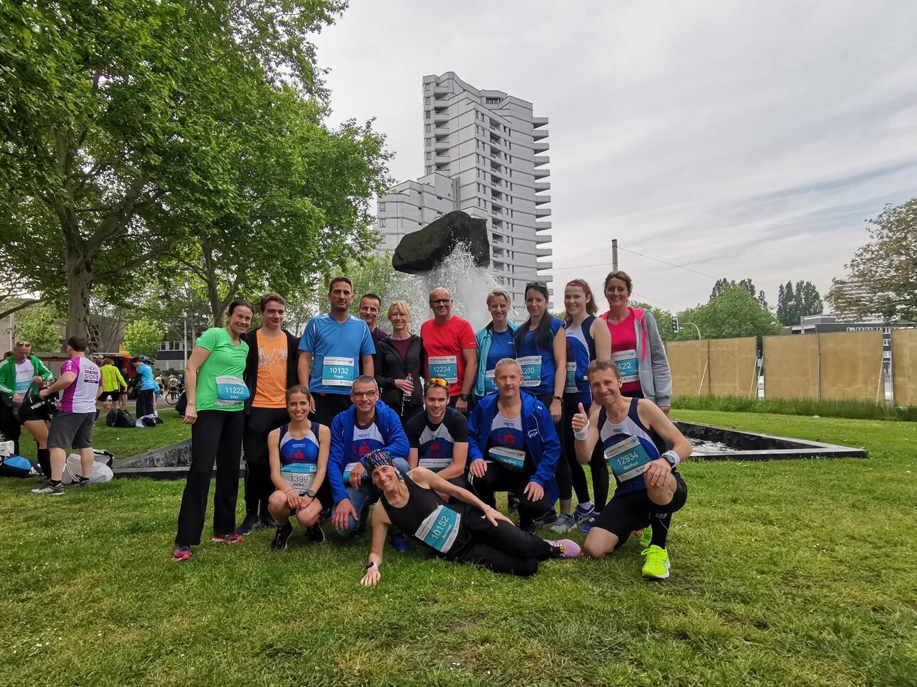 Gruppe von Läufern mit Startnummern steht neben einem Wasserfall in einem Park, umgeben von Bäumen und modernen Gebäuden.