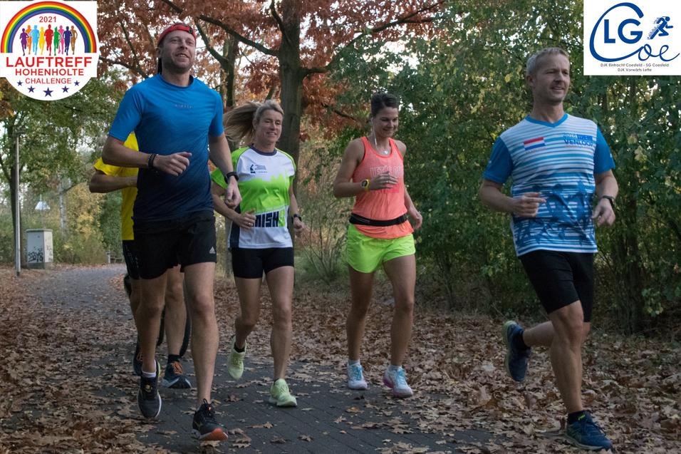 Gruppe von fünf Läufern in Sportkleidung joggt auf einem mit Herbstlaub bedeckten Weg durch einen Park.