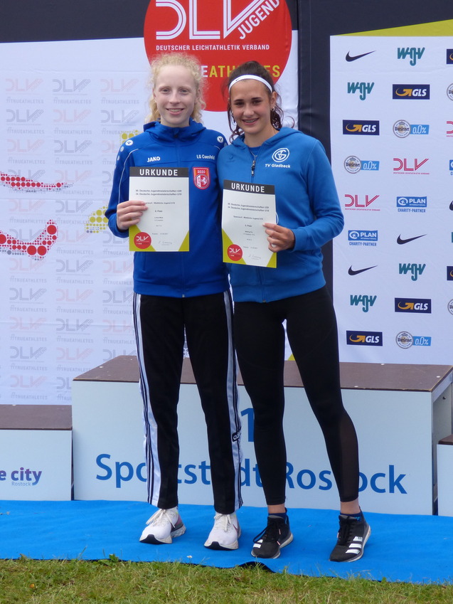 Zwei junge Athletinnen auf dem Podium halten Urkunden in der Hand, mit Sponsorenlogos im Hintergrund.