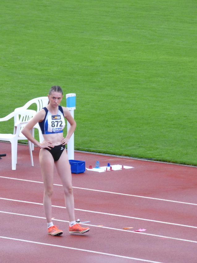 Athletin mit Startnummer 872 steht auf einer Laufbahn und wartet auf den Wettkampf, im Hintergrund eine grüne Rasenfläche.