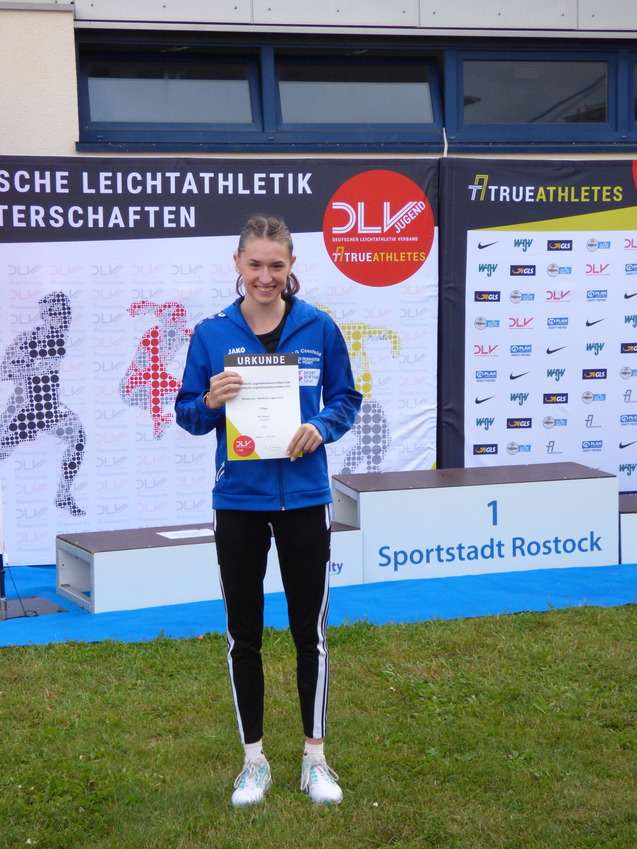 Junge Sportlerin in blauer Jacke hält Urkunde vor dem Banner der Deutschen Leichtathletikmeisterschaften.
