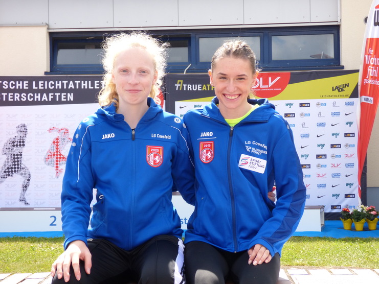 Zwei Athletinnen in blauen Trainingsjacken der LG Coesfeld posieren lächelnd vor einem Event-Hintergrund.