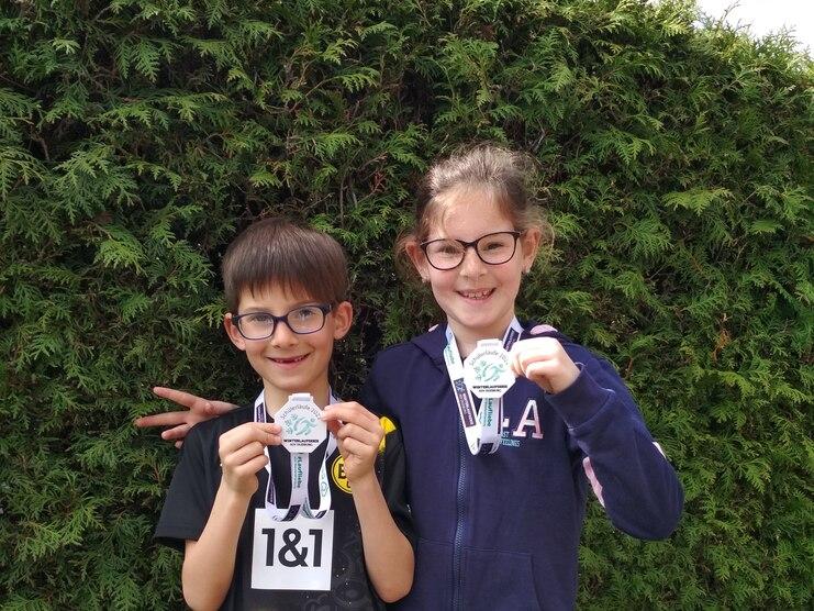 Zwei Kinder halten Medaillen in der Hand, l&auml;cheln vor einer gr&uuml;nen Hecke und tragen Sportkleidung mit Nummern.