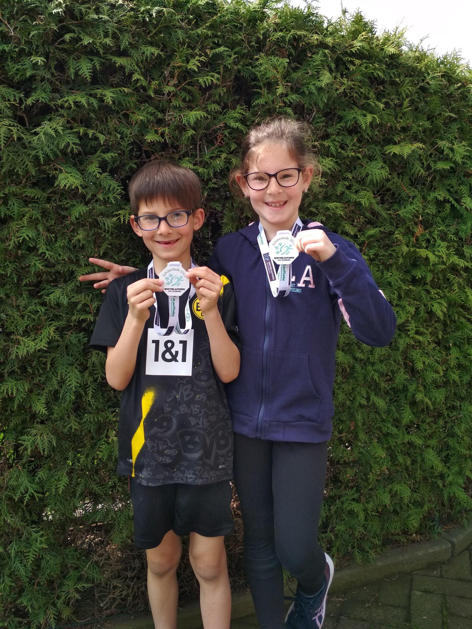 Zwei Kinder halten Medaillen in der Hand, lächeln vor einer grünen Hecke und tragen Sportkleidung mit Nummern.