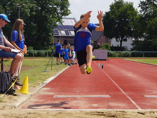 Ein Junge springt im Weitsprung über eine Sandschicht auf einer roten Laufbahn, Zuschauer beobachten im Hintergrund.