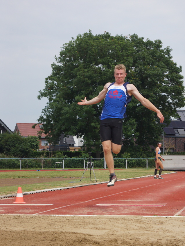 Ein Hochspringer in Aktion, der in den Sandbereich eines Sprungrohrs springt, im Hintergrund eine weitere Athletin.