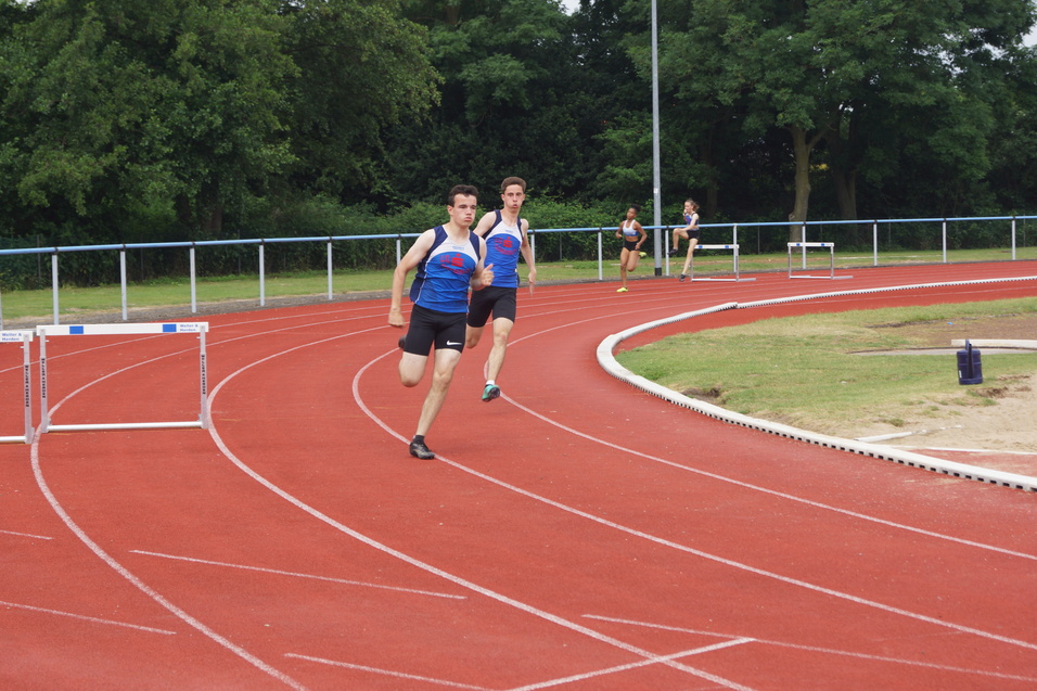 Zwei Athleten sprinten auf einer roten Laufbahn, während ein weiterer Sportler in der Nähe trainiert.