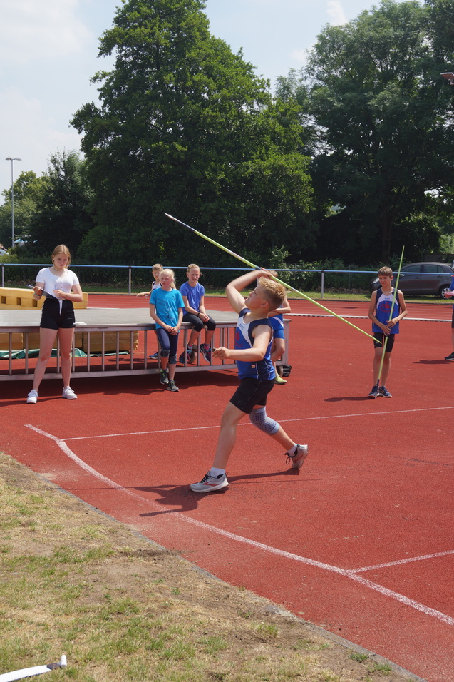 Ein Junge wirft einen Speer auf einem roten Sportplatz, während andere Kinder ihn beobachten.