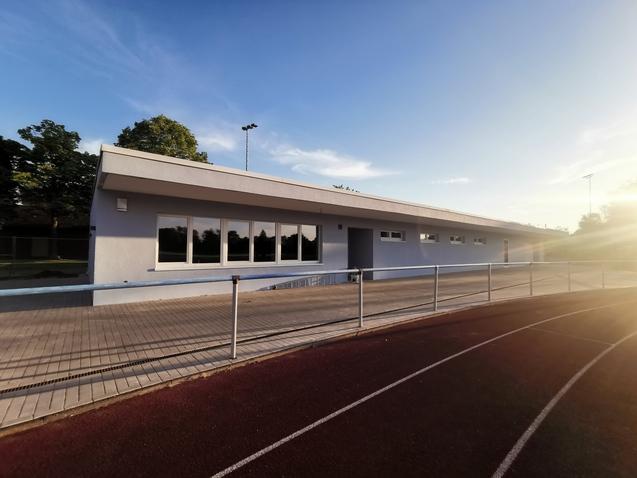 Moderne Sporteinrichtung mit großen Fenstern, Terrasse und Laufbahn im Vordergrund an einem klaren Himmel.