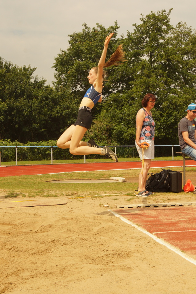 Athletin springt im Weitsprung über eine Sandgrube, Zuschauer stehen im Hintergrund auf einem Sportplatz.