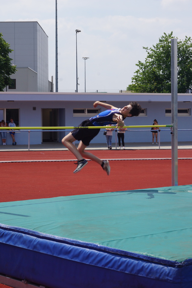 Ein Hochspringer springt über eine gelbe Latte auf einer roten Laufbahn, hinter ihm Zuschauer und ein Sportgebäude.