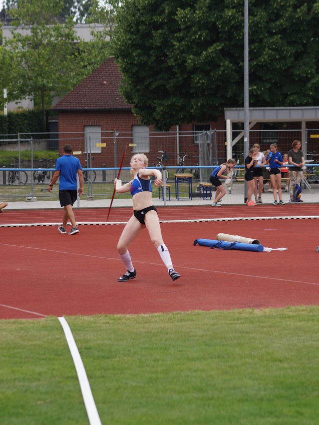Athletin mit einem Speer in der Wurfauslage auf einem roten Sportplatz, Hintergrund: Zuschauer und weitere Athleten.