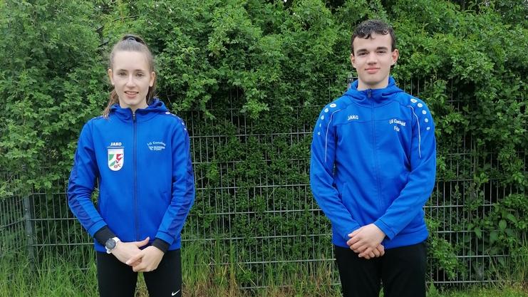 Zwei Jugendliche in blauen Trainingsjacken stehen im Freien vor gr&uuml;ner Hecke auf einer Wiese.