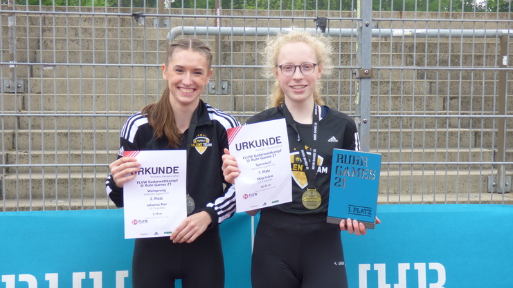 Zwei junge Athletinnen halten ihre Urkunden und Trophäen vom Ruhr Games 21, lächelnd vor einem blauen Hintergrund.