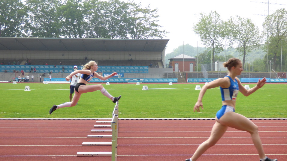 Zwei Athletinnen sprinten über eine Hürde auf einer Leichtathletikbahn. Zuschauer sind im Hintergrund sichtbar.