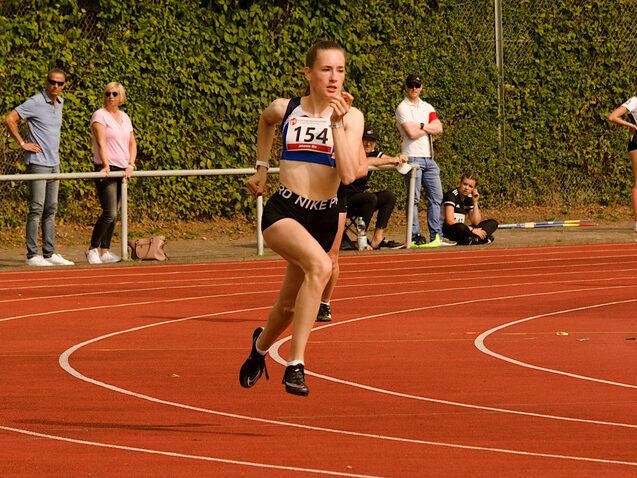 Eine L&auml;uferin in Sportbekleidung sprintet auf einer roten Laufbahn, Zuschauer stehen im Hintergrund.