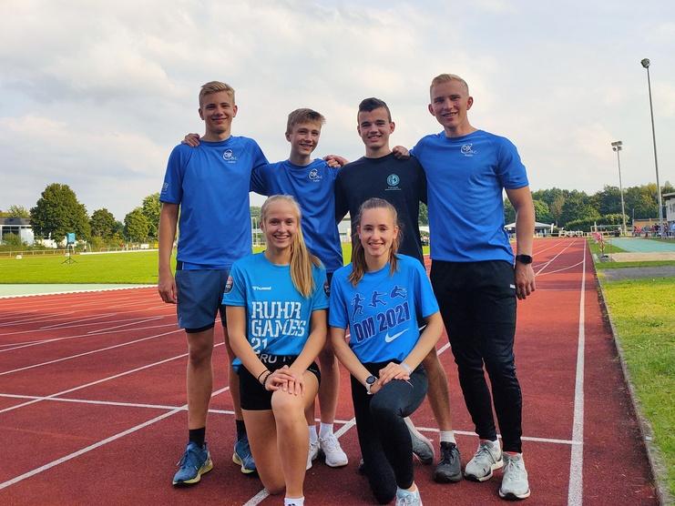 Sechs Sportler in blauen T-Shirts posieren auf einer Laufbahn, mit einem Sportgel&auml;nde im Hintergrund.