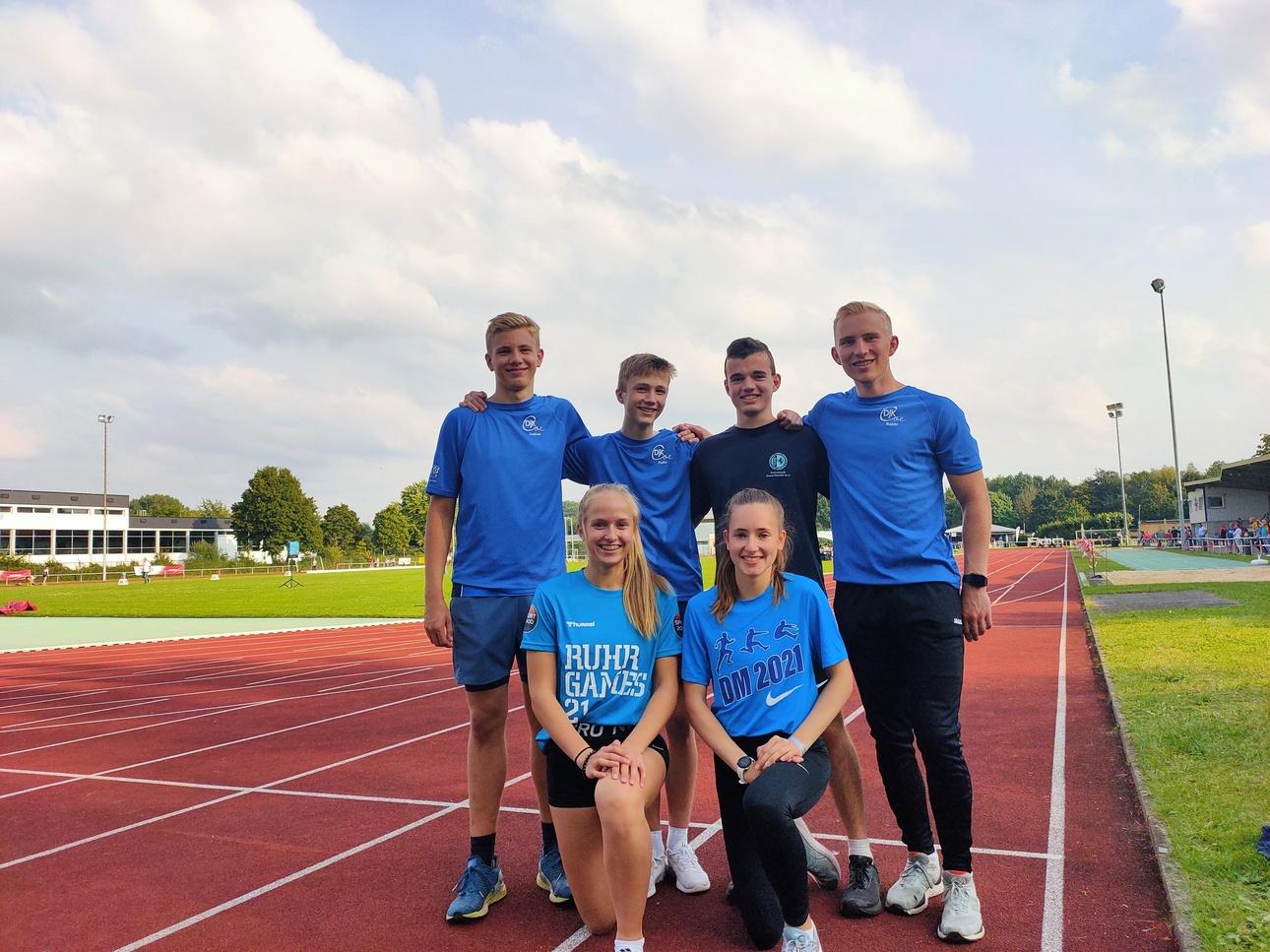 Sechs Sportler in blauen T-Shirts posieren auf einer Laufbahn, mit einem Sportgelände im Hintergrund.