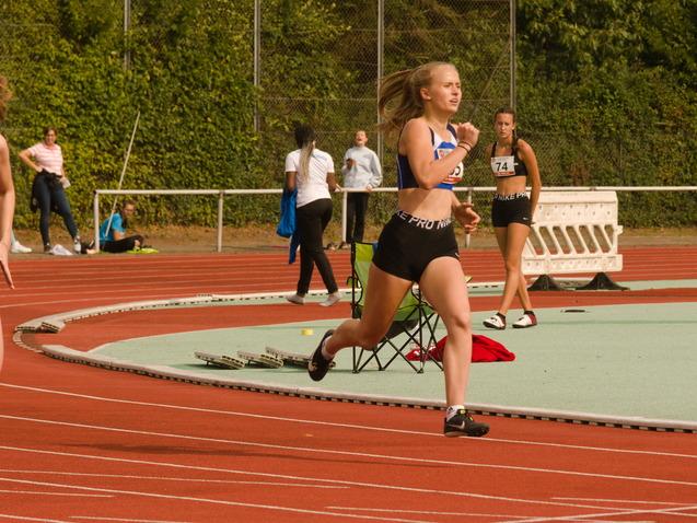 Zwei Athletinnen laufen auf einer Laufbahn während eines Wettkampfs; Zuschauer und weitere Teilnehmer im Hintergrund.