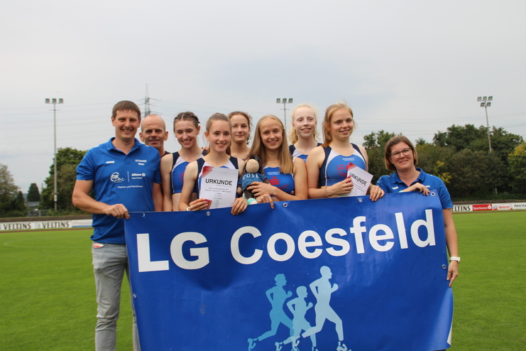Gruppe von Athleten und Trainern der LG Coesfeld mit einem Banner auf einem Sportplatz, einige halten Urkunden und einen Teddy.