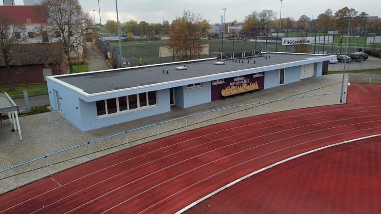 Blick auf ein Sportheim mit blauer Außenwand, neben einem roten Laufbahnring und Fußballfeldern im Hintergrund.