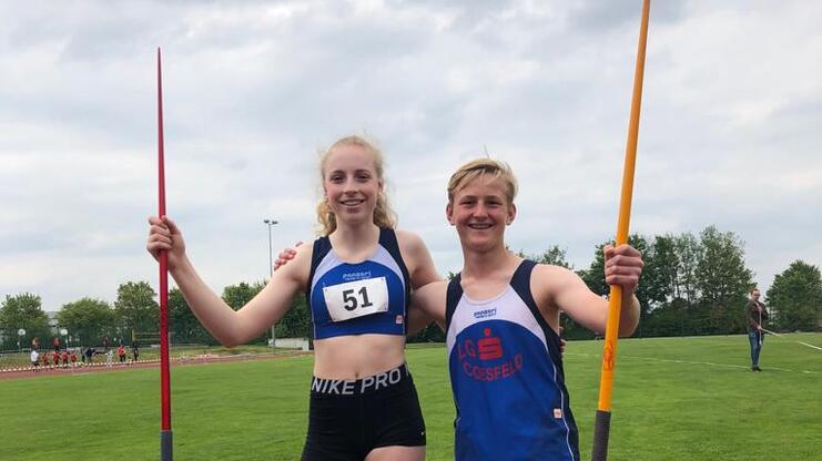 Zwei Jugendliche stehen auf einem Sportplatz, jeder mit einem Speer in der Hand, neben einer Laufbahn.
