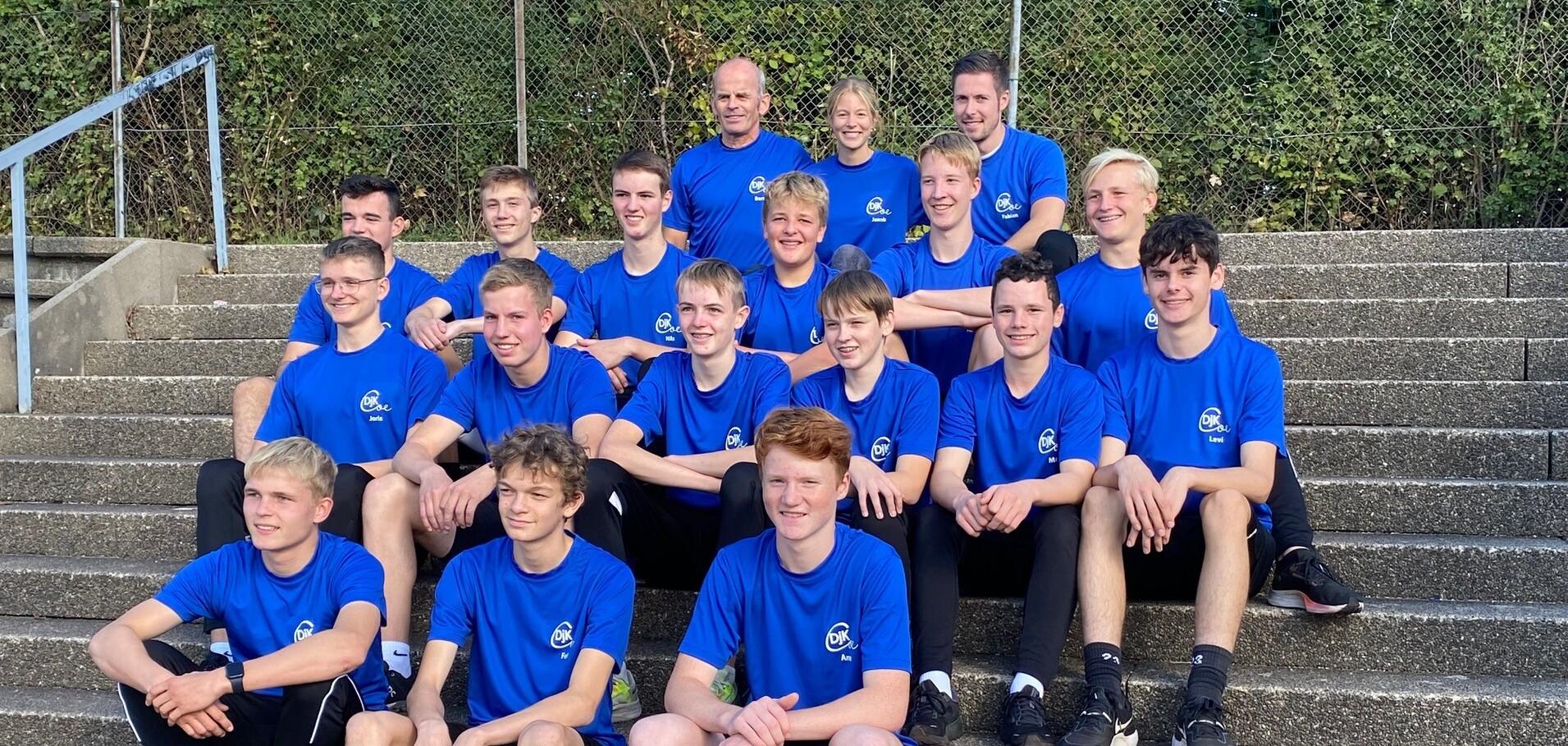 Gruppe von Jugendlichen in blauen T-Shirts sitzt auf einer Treppe, im Hintergrund sind Bäume sichtbar.
