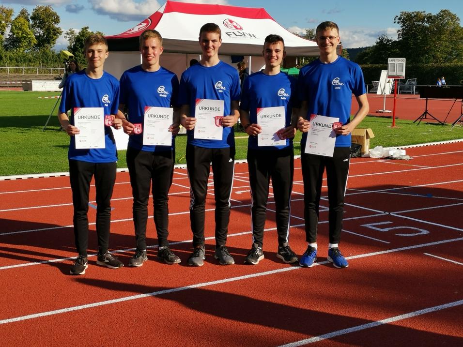 Fünf Leichtathleten in blauen T-Shirts halten Urkunden auf einer Rennbahn unter einem Zelt.