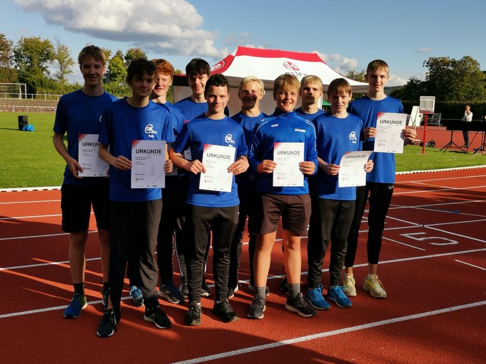 Gruppe von jungen Sportlern in blauen T-Shirts, die stolz Urkunden auf einer Laufbahn halten.