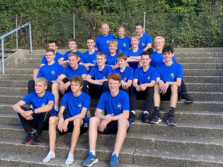 Gruppe von Jugendlichen in blauen T-Shirts sitzt auf einer Treppe, im Hintergrund sind Bäume sichtbar.