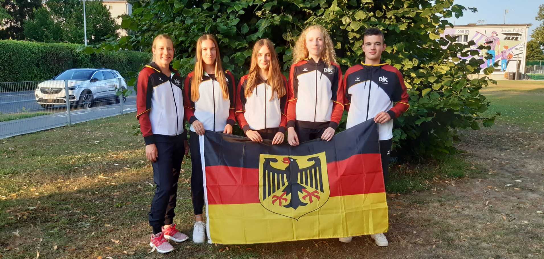 Fünf Personen stehen im Freien und halten eine deutsche Flagge vor einem Baum. Sie tragen Trainingsjacken.