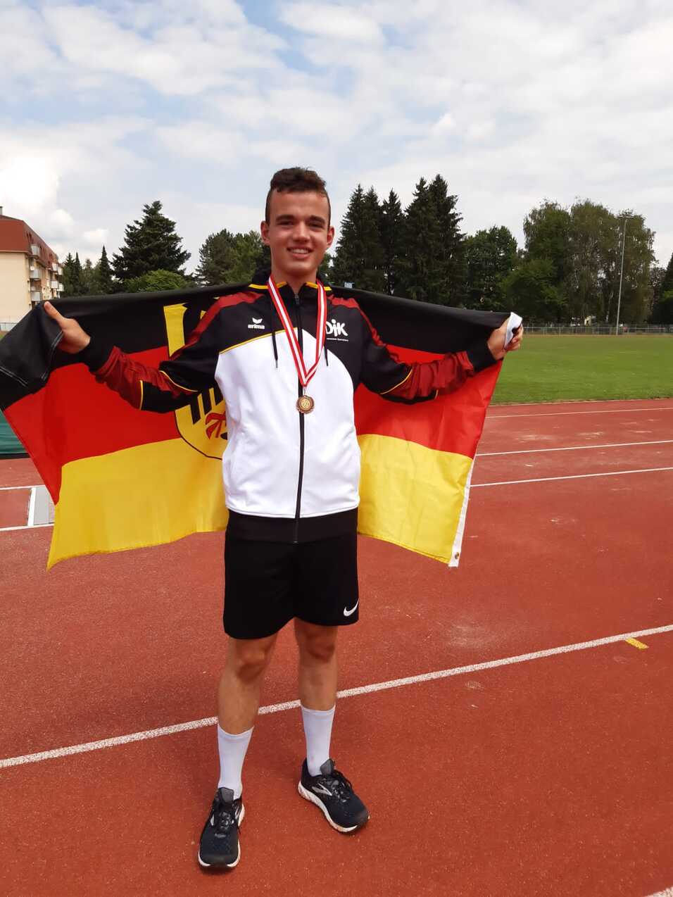 Junger Athlet hält eine deutsche Fahne und trägt Medaillen auf einem Sportplatz. Hintergrund mit Bäumen und Stadien.