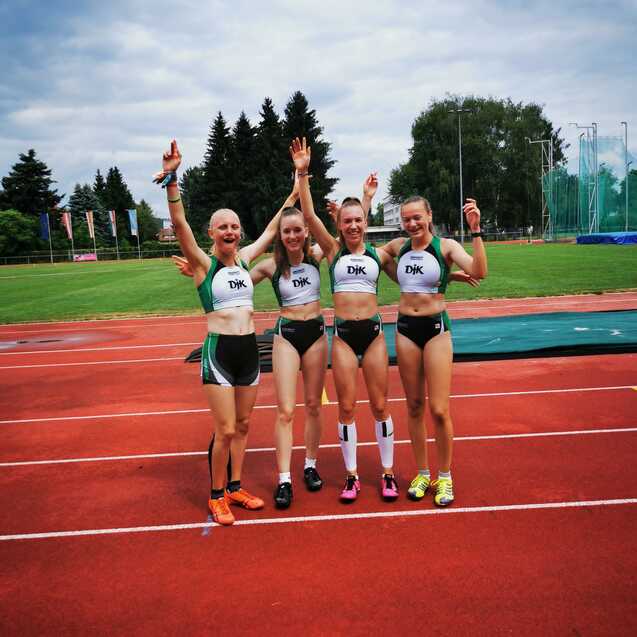 Vier feiernde Athletinnen stehen auf einer Tartanbahn, tragen Sportbekleidung und zeigen ihre Erfolge mit erhobenen Händen.