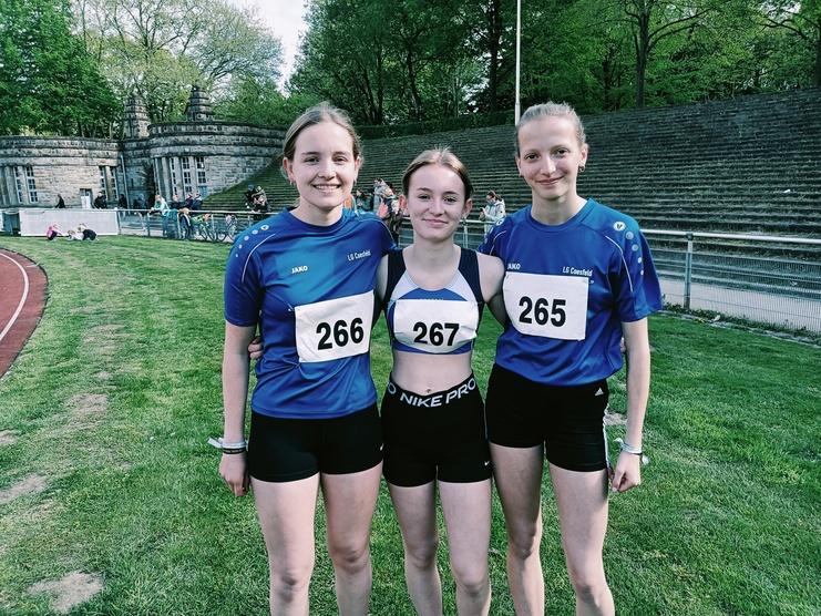 Drei Läuferinnen in Sportbekleidung mit Startnummern stehen auf einer Laufbahn vor einem sportlichen Hintergrund.