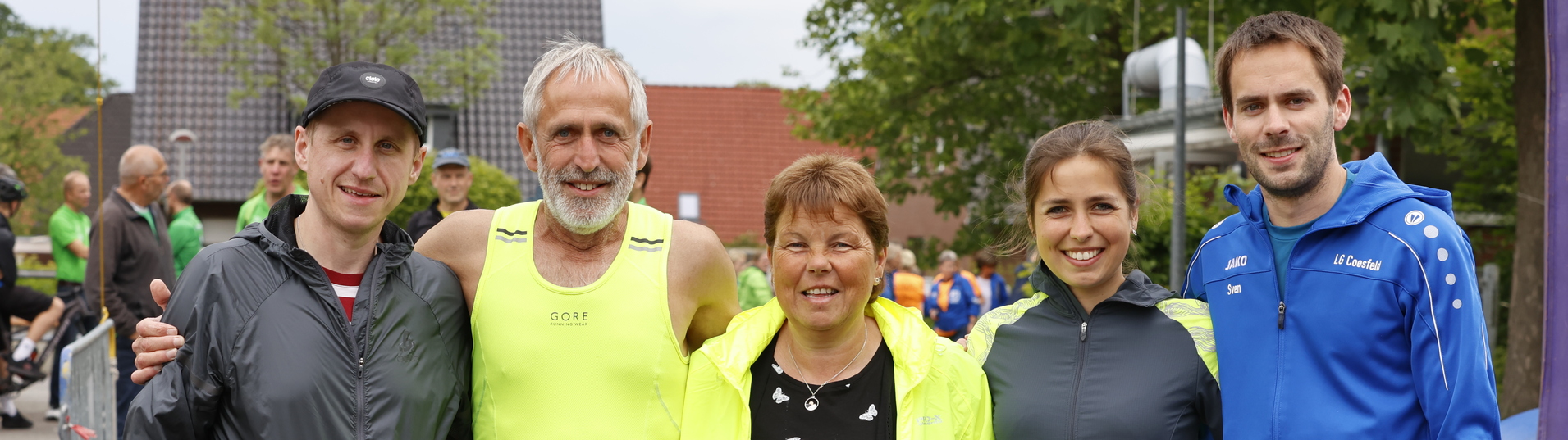 Fünf Personen in Sportbekleidung stehen lächelnd nebeneinander vor einer Laufveranstaltung im Freien.