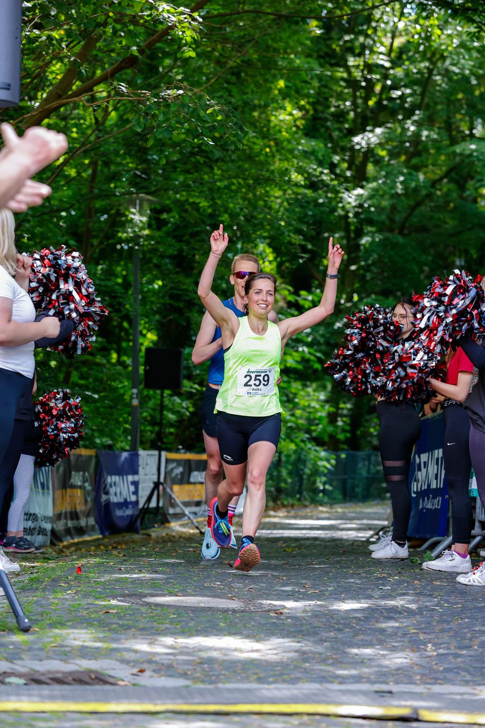 Läuferin mit Startnummer 259 jubelt beim Zieleinlauf, umgeben von jubelnden Zuschauern und Cheerleadern im Wald.