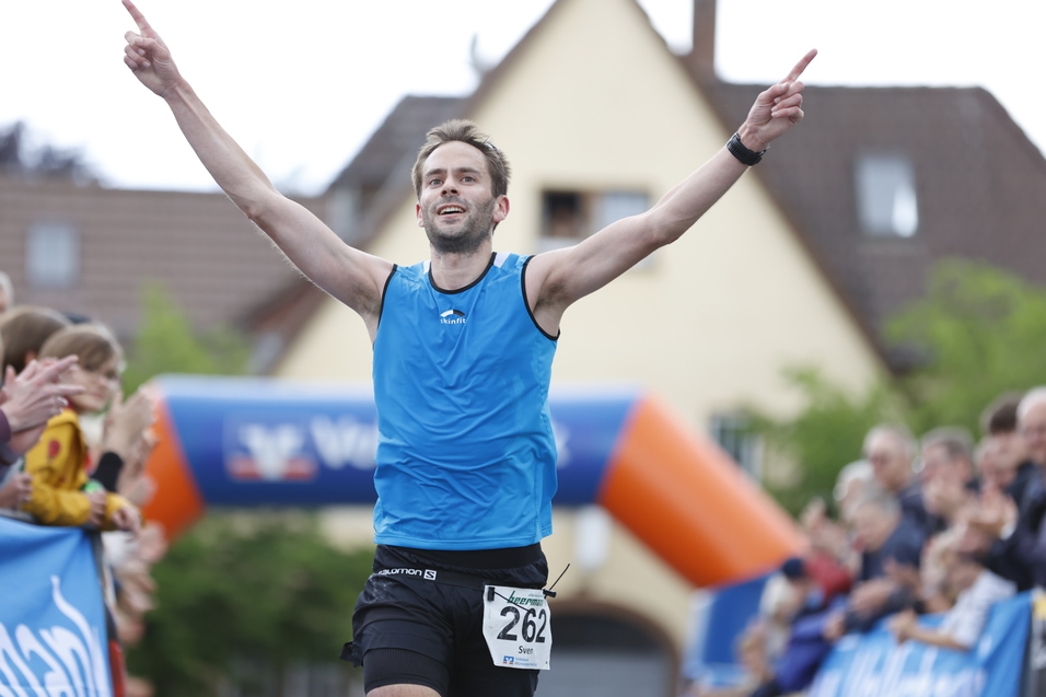 Mann in blauer Sportbekleidung jubelt nach dem Ziel eines Rennens, umgeben von applaudierenden Zuschauern.