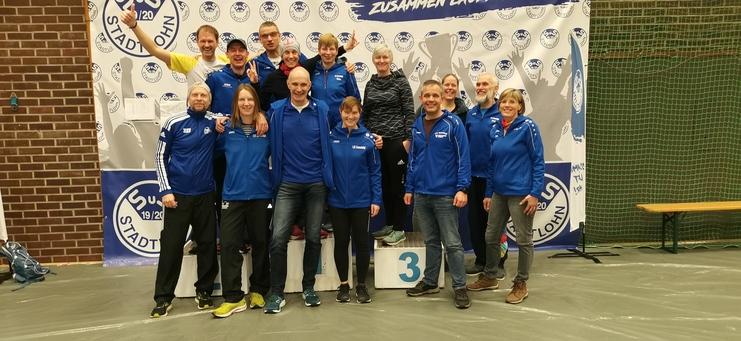 Gruppe von Teilnehmern in blauen Sportjacken posiert fröhlich auf einem Siegerpodest vor einem Banner.