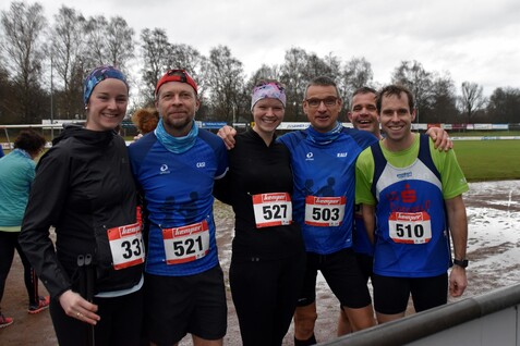 Sechs laufbegeisterte Personen posieren mit Startnummern an einem Sportfeld, umgeben von nassem Boden und Bäumen.