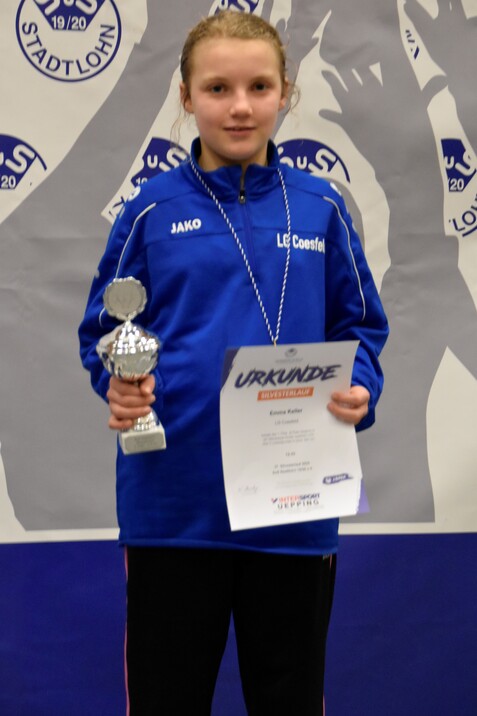 Mädchen in blauem Sportanzug hält Pokal und Urkunde für den Silvesterlauf in der Hand, vor einem gemusterten Hintergrund.