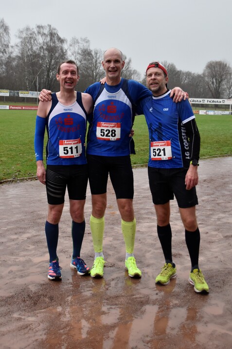 Drei Läufer in Sportbekleidung stehen lächelnd auf einer nassen Laufstrecke, jeder trägt eine Startnummer.