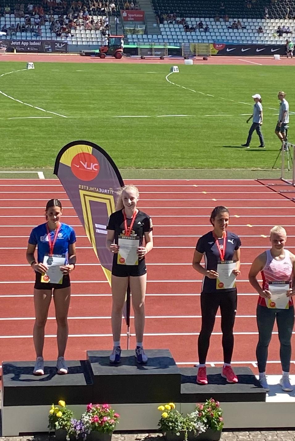 Vier Athletinnen stehen auf Podesten mit Urkunden und Medaillen auf einer Leichtathletikanlage unter blauem Himmel.