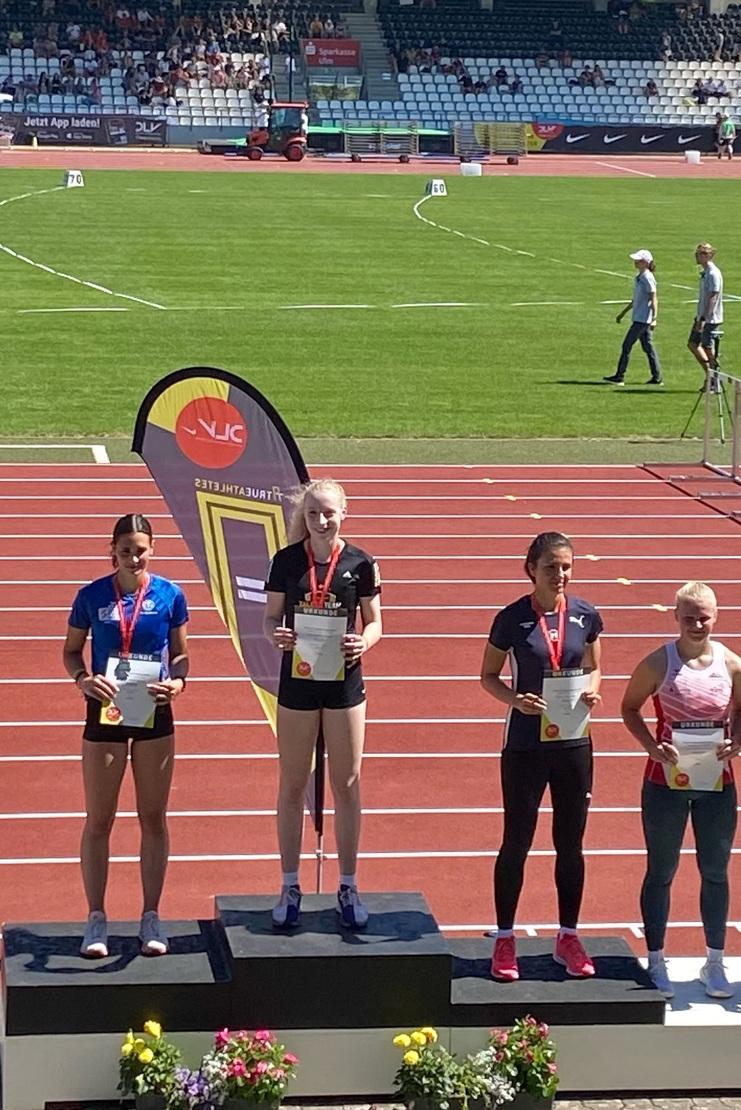 Vier Athletinnen stehen auf Podesten mit Urkunden und Medaillen auf einer Leichtathletikanlage unter blauem Himmel.