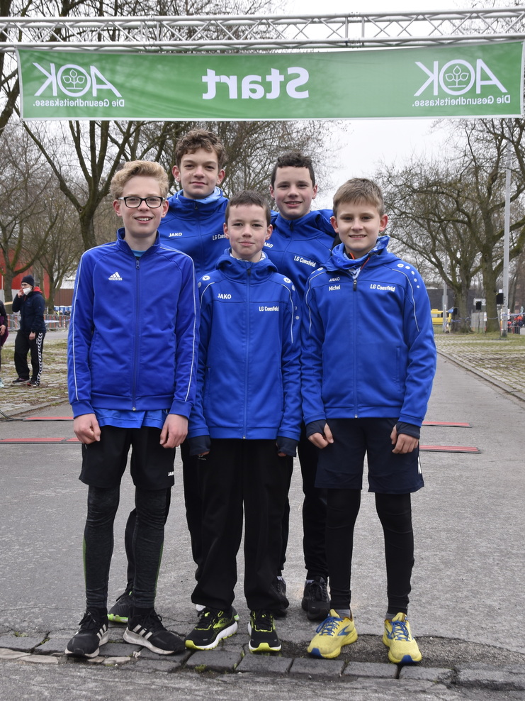 Fünf Jugendathleten in blauen Trainingsjacken stehen am Startbogen eines Laufwettbewerbs im Freien.