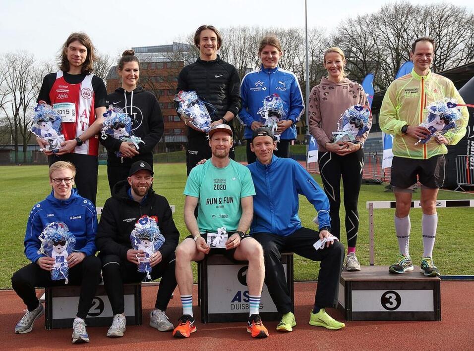 Gruppenbild von Laufteilnehmern auf dem Siegerpodest mit Blumensträußen, im Hintergrund ein Sportplatz.