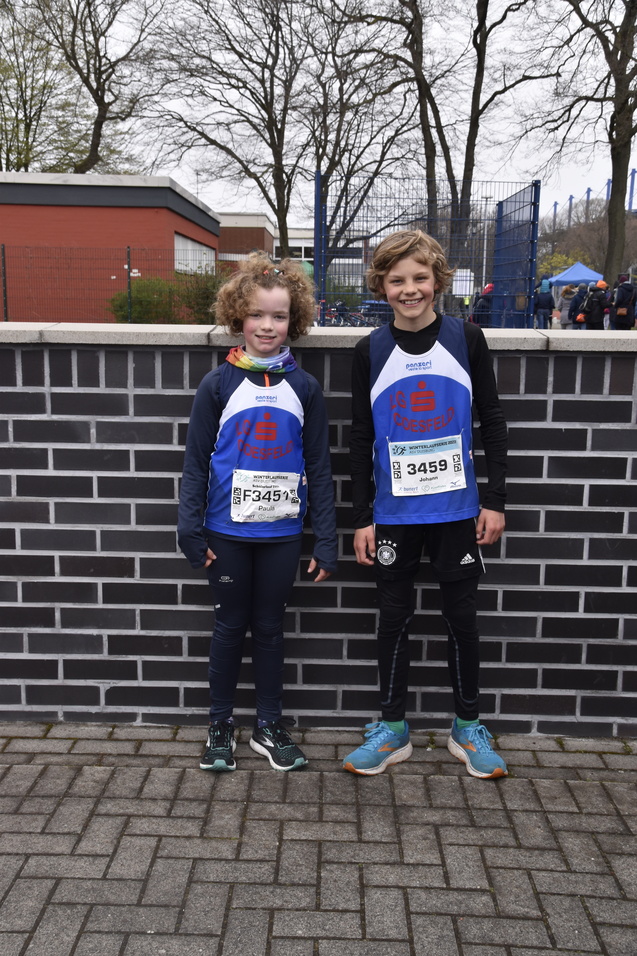 Zwei Kinder in Laufkleidung stehen vor einer Mauer, bereiten sich auf ein Rennen vor und halten ihre Startnummern.