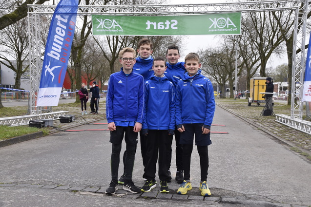 Fünf Jugendathleten in blauen Trainingsjacken stehen am Startbogen eines Laufwettbewerbs im Freien.