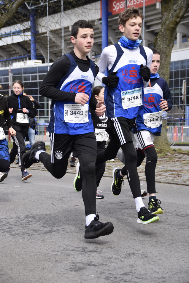 Vier junge Läufer in Sportkleidung rennen auf einer Straße, einige tragen Startnummern. Im Hintergrund sind Zuschauer zu sehen.